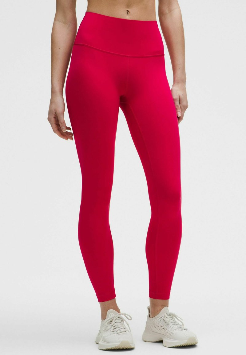 Rde hlače visokega pasu, izdelane iz elastičnega materiala, z gladko teksturo in oprijetim dizajnom, v kombinaciji z belimi športnimi čevlji.