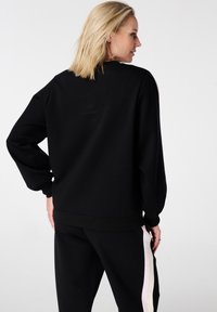 Zwart sweatshirt met lange mouwen, geribbelde manchetten en zoom. Zachte textuur met een ontspannen pasvorm, gedragen met bijpassende zwarte broek met strepen aan de zijkant.