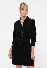 Svart kordfløyelskjole med lange ermer, krage, frontlukking med knapper, belte i midjen og sidelommer. Myk tekstur og tettsittende design.