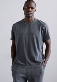 Tricou din bumbac gri cu mâneci scurte, guler rotund și un design de text subtile. Textura moale și croiala lejeră completează pantalonii gri asortați.