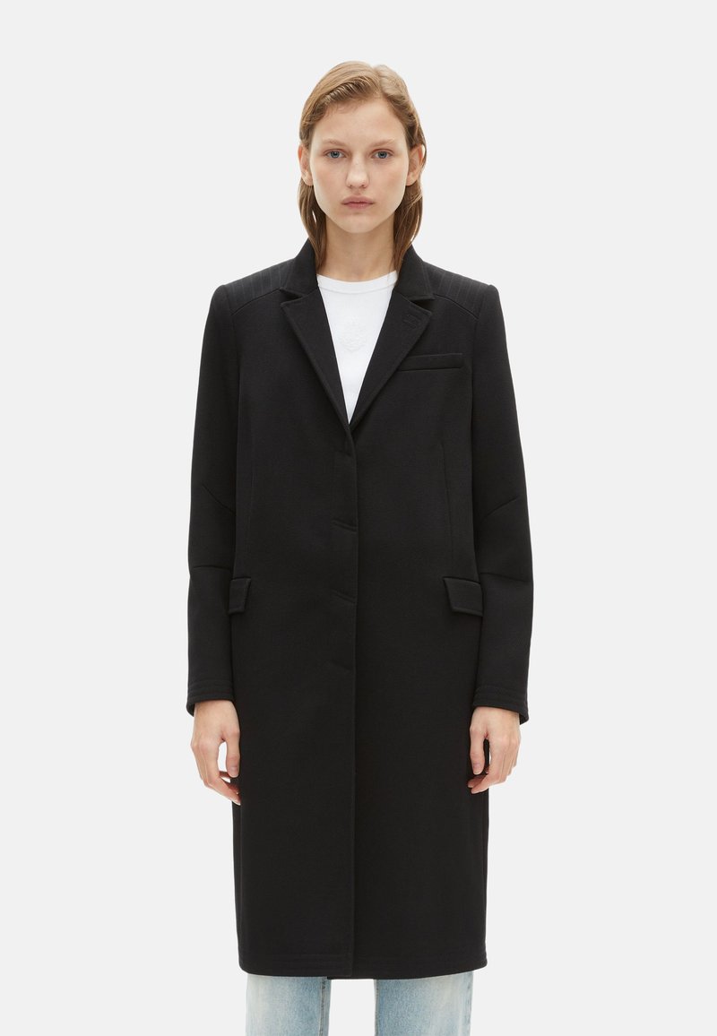 Manteau noir ajusté avec un col cranté, des manches longues, un tissu texturé, des poches latérales et une coupe droite, porté sur un haut blanc.