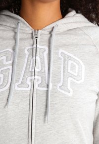 Szara bluza z kapturem zapinana na zamek, wykonana z miękkiego materiału, z dużym białym napisem "GAP" na przodzie, posiadająca kaptur ze sznurkiem i gładką fakturę.