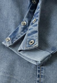 Gros plan sur un tissu en denim bleu clair avec trois boutons-pression en métal partiellement fixés sur un bord plié.
