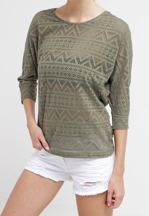 Vrouw draagt een doorschijnende, olijfgroene, langmouwige top met patroon en witte denim shorts met scheuren, staat met één hand in de zak.