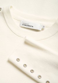 Kreemvärvi kudum, millel on soonikkoes kaelus ja mansetid, ning hõbedased silmade detailid varrukatel. Sildil on kirjas "Karen by Simonsen," suurus M.