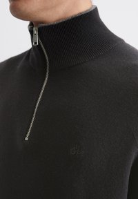 Čierny sveter na zips s vysokým žebrovaným golierom, ktorý má strieborný zips a diskrétny detail loga. Vyrobený z mäkkého, textúrovaného materiálu.
