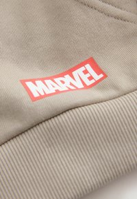 Beige sviiterikangas sisaldab ribikanti ja silmatorkavat punast ristkülikukujulist "MARVEL" logo valge tekstiga.