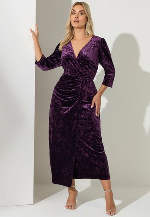 Kvinna klädd i en lång lila sammets-wrap-klänning med rynkade detaljer, stående med ena handen på en vägg och iförd silvriga högklackade sandaler.