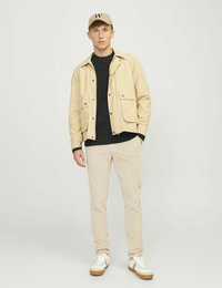 Beige pamut dzseki két elülső zsebbel, fekete kötött pulóver fölött, világos beige nadrággal és barna talpú fehér sportcipővel párosítva.