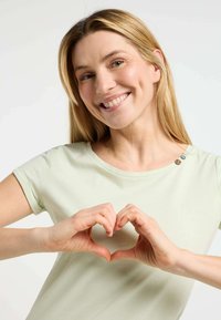 Camicia verde chiaro a maniche corte con scollo rotondo e bottoni decorativi sulla spalla. Mani che formano un gesto a cuore davanti.