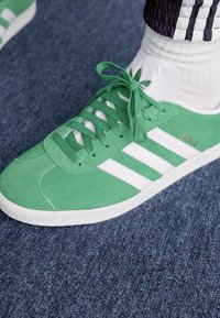 Green suede sneaker with white three-stripe design, flat rubber sole, and round toe. Laces in matching green. Model name "Gazelle" featured.