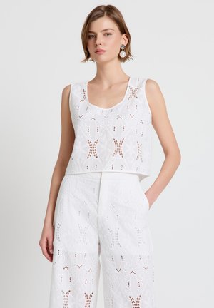 Donna che indossa un top corto in pizzo bianco senza maniche e pantaloni a gamba larga a vita alta coordinati, con orecchini a goccia di perle, in piedi davanti a uno sfondo semplice.