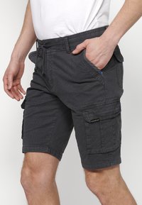 Man i mörkgrå cargoshorts med flera fickor och en vit t-shirt, hand i framfickan mot en enkel bakgrund.