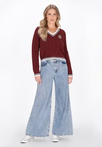 Pulover tricotat maroon cu decolteu în V, dungi albe contrastante și emblema. Asortat cu blugi largi, de culoare deschisă, și adidași albi.
