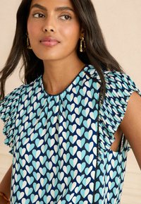 Blå mönstrad topp som har ljusblå och vita hjärtformer, med volangärmar och rund halsringning. Lättviktsmaterial med en mjuk textur.