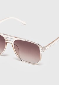 Klare Brillengestell-Sonnenbrille mit verlaufenden dunklen Gläsern und schlanken transparenten Bügeln auf einer hellgrauen Oberfläche.