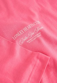 Roza bombažna majica z belim napisom: "Lonely Hearts Club, Dolce Far Niente," z gladko teksturo in standardnim krojem.