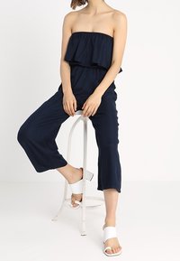 Žena sedící na bílém kovovém stoličce, oblečená v námořnickém modrém overal bez ramínek a bílých sandálech na blokovém podpatku.