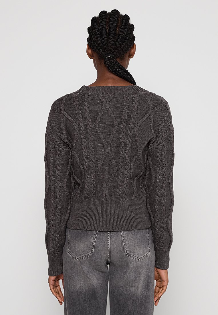 Persoon met gevlochten haar, gekleed in een donkergrijze kabeltrui en vervaagde grijze jeans, met zijn rug naar de camera tegen een effen achtergrond.