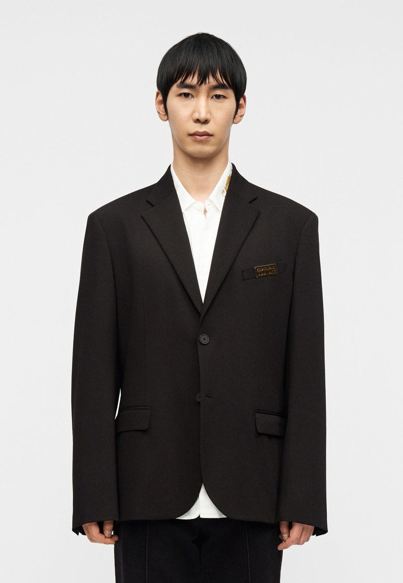 Νεαρός άνδρας με ίσια μαύρα μαλλιά, φορώντας ένα φαρδύ μαύρο σακάκι πάνω από λευκό πουκάμισο, στέκεται μπροστά σε ένα απλό φωτεινό φόντο.