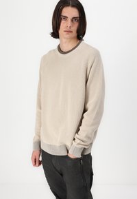 Suéter de punto acanalado beige con cuello redondo y detalles grises en los puños. Combinado con pantalones oscuros, mostrando un ajuste relajado.