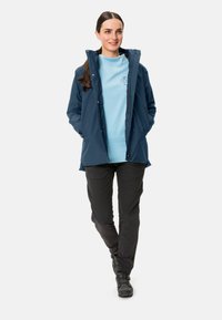 Giacca impermeabile blu navy con colletto alto, abbinata a una felpa azzurro chiaro e pantaloni neri slim-fit, completata da scarpe nere.
