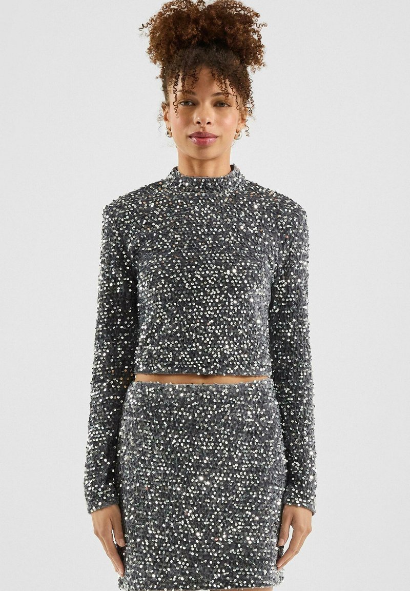 Silberner, mit Pailletten besetzter Cropped-Pullover mit langen Ärmeln, kombiniert mit einem passenden, hochgeschnittenen Rock. Glatte Textur mit reflektierenden Elementen.