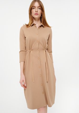 Tansfarbene Knopfhemdbluse mit Kragen, Dreiviertelärmeln, verstellbarem Kordelzug in der Taille und knielanger Länge. Hergestellt aus glattem Stoff.