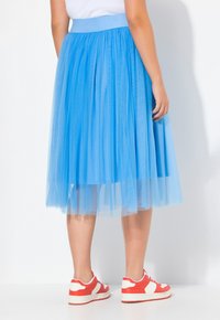 Gonna in tulle blu con una cintura larga, design a strati e texture trasparente. Abbinata a sneaker rosse e bianche su uno sfondo semplice.