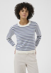 Femme aux cheveux bouclés portant un pull rayé bleu et blanc et un pantalon beige clair, debout avec les mains derrière le dos.