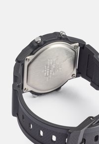 Orologio con cinturino in gomma nera a finitura texturizzata, fondello in acciaio inossidabile e due pulsanti. Presenta testo inciso sulla superficie metallica.