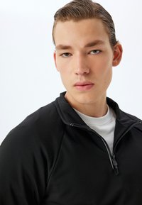 Pulóver negro de media cremallera en tejido suave con cuello alto. Llevado sobre una camiseta blanca, con cremallera y detalles de costura sutiles.