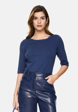 Woman wearing a navy blue three-quarter sleeve top and high-waisted dark blue patterned leather pants, posing with one hand in pocket.