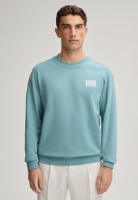 Felpa azzurra chiaro realizzata in morbido cotone, con scollatura rotonda, polsini a coste e un piccolo logo bianco sul lato sinistro del petto.