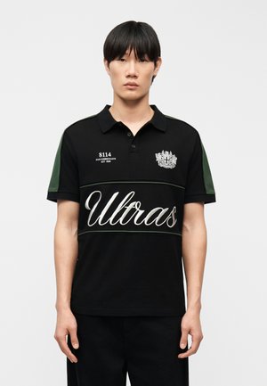 Mees, kes kannab musta lühikeste varrukatega polo särki, millel on kirjad "Ultras", "8114" ja vappemblem, seisab ühetaolise heleda tausta ees.