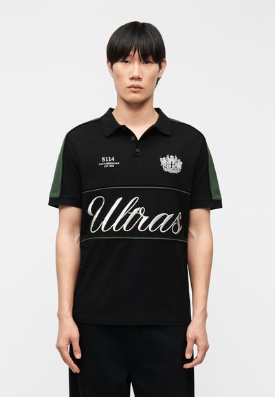 Homme portant un polo noir à manches courtes avec les inscriptions « Ultras », « 8114 » et un écusson, debout devant un fond clair uni.