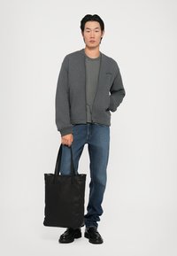 Hanorac gri cu fermoar peste un tricou închis, blugi, geantă de umăr neagră cu două bretele și pantofi negri. Design simplu, stil casual.