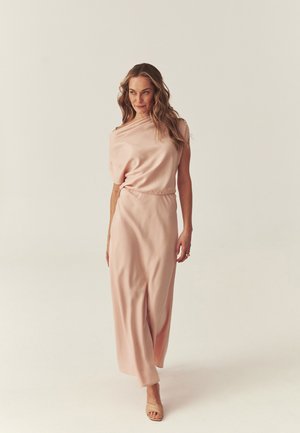 Woman walking in a pale pink, one-shoulder satin dress with a floor-length skirt and beige open-toe heels against a plain background.