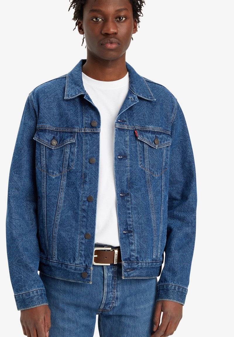 Veste en denim bleu foncé, avec des fermetures à boutons, deux poches poitrine et un tissu légèrement texturé. Portée sur un t-shirt blanc.