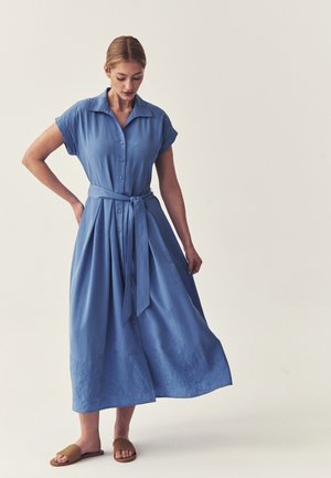 Donna che indossa un abito midi blu con bottoni, maniche corte e cintura in vita, in piedi a piedi nudi su uno sfondo chiaro e uniforme.