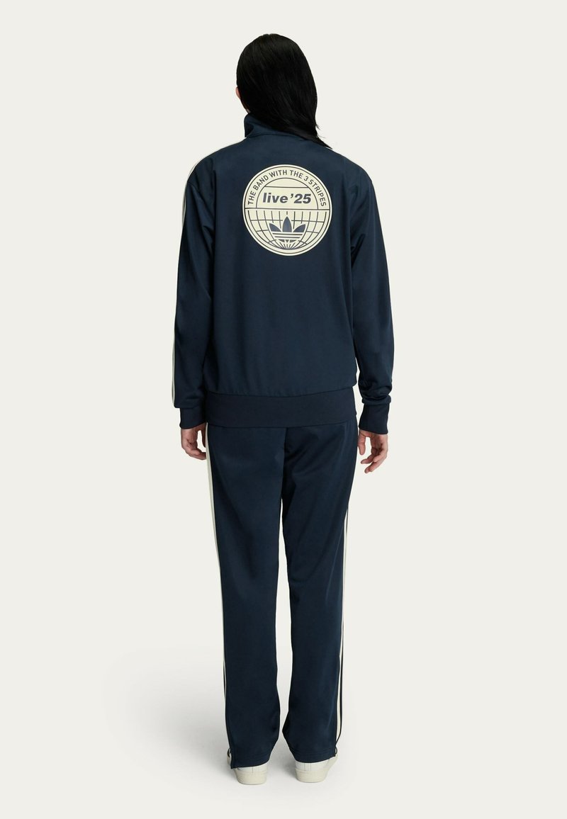 Navy tracksuit features a high-neck jacket with a circular logo at the back and white side stripes on the pants. Made of smooth fabric.