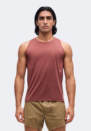 Mann mit dunklem Haar trägt ärmelloses bordeauxfarbenes Sportoberteil und beige Shorts, steht frontal vor einfarbigem Hintergrund.