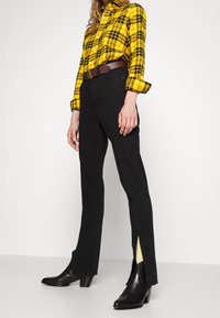 Donna che indossa una camicia a scacchi gialla infilata in pantaloni neri a vita alta con fessure laterali e stivaletti neri contro uno sfondo bianco.