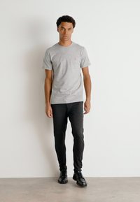 Camiseta gris de manga corta con un pequeño logotipo en el pecho, combinada con pantalones negros ajustados y zapatos negros de cordones. Fondo liso.