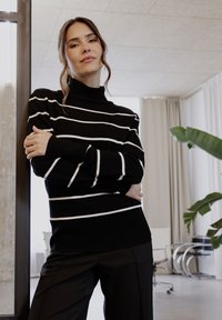 Schwarz-weißer gestreifter Rollkragenpullover mit langen Ärmeln, kombiniert mit schwarzen Hosen. Glattes Material, bequemer Schnitt und gerippte Details an den Bündchen.