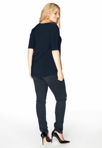 Marineblå, kortærmet top i blødt stof, parret med mørke jeans. Modellen bærer sorte højhælede sko, som viser et afslappet look fra bagsiden.