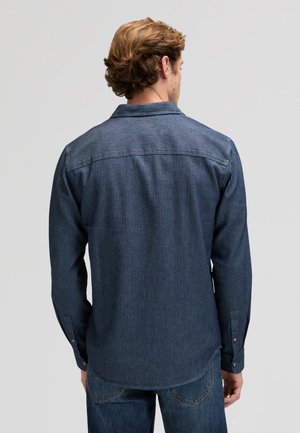 Moški s svetlo rjavimi lasmi, oblečen v temno modro majico z dolgimi rokavi s črtami in modre kavbojke, prikazan od zadaj na enobarvnem ozadju.