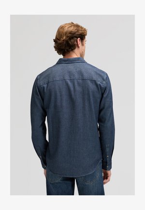 Mees helepruunide juustega, kes kannab tumesinist triibulist pikade varrukatega särki ja siniseid teksapükse, nähtud tagantpoolt tavalise tausta ees.