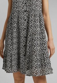 Femme portant une robe noire mi-longue à motif floral blanc avec des boutons noirs sur le devant, les bras détendus le long du corps.
