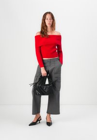 Pull en maille rouge à épaules dénudées, pantalon cargo large gris et sac à main texturé noir. Escarpins noirs à bout pointu et à bride arrière.